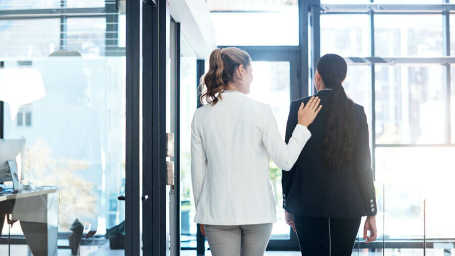 Two coworkers stand with one putting her hand on the back of the other as they decide to take action.