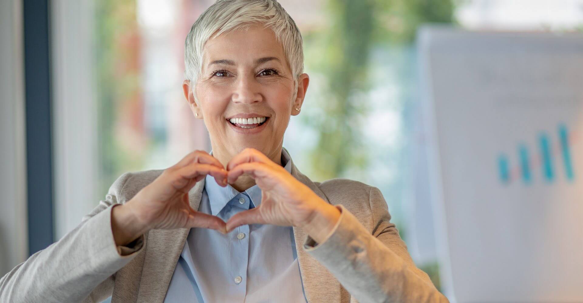 Portrait of beautiful mature woman in a business office shows heart with her hands, connecting investments to the heart