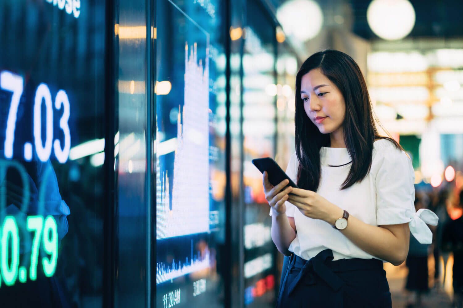 Confident young Asian businesswoman checking financial trading data on smartphone by the stock exchange market display screen board in downtown financial district