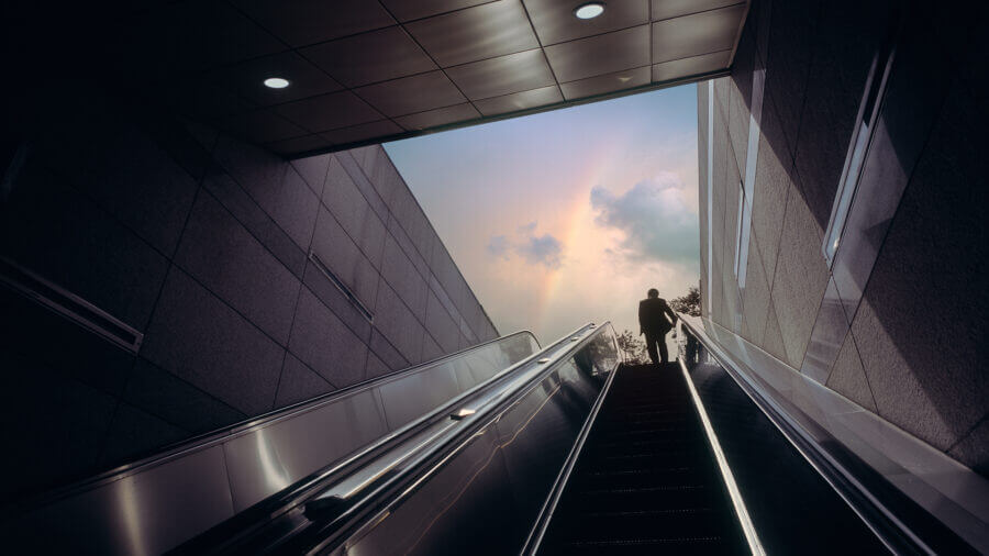 Escalator of subway station with businessman moving towards dramatic sky with rainbow.