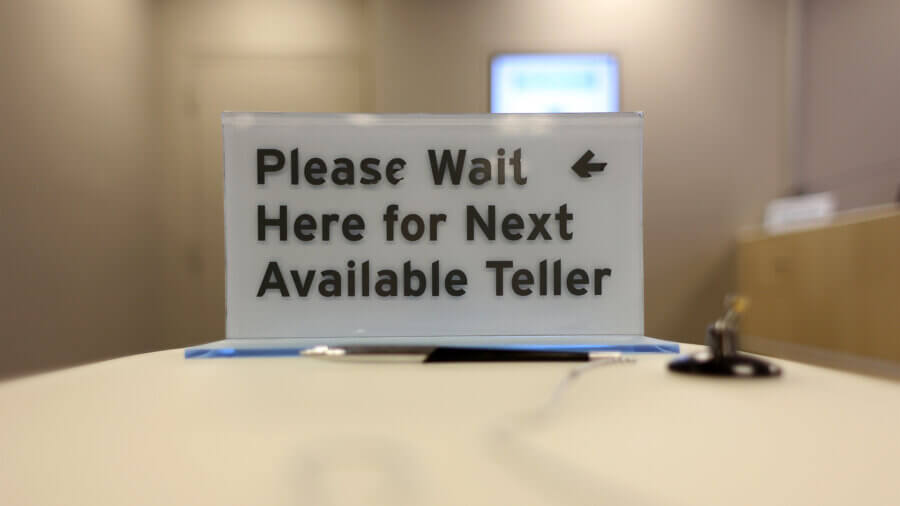 A sign telling bank customers to wait for the next available bank teller.