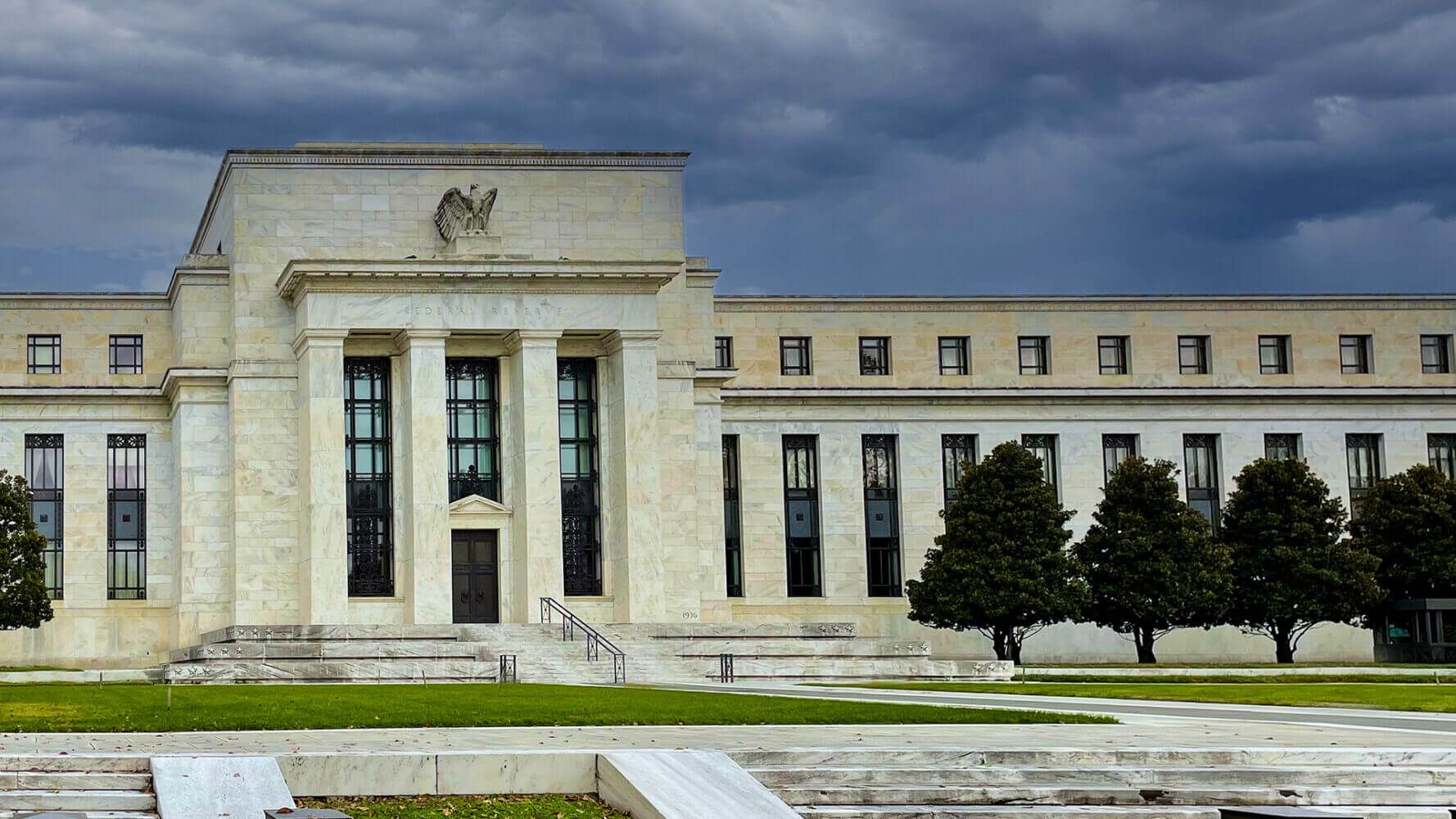 The Federal Reserve building stands tall and stark in contrast to a stormy background.