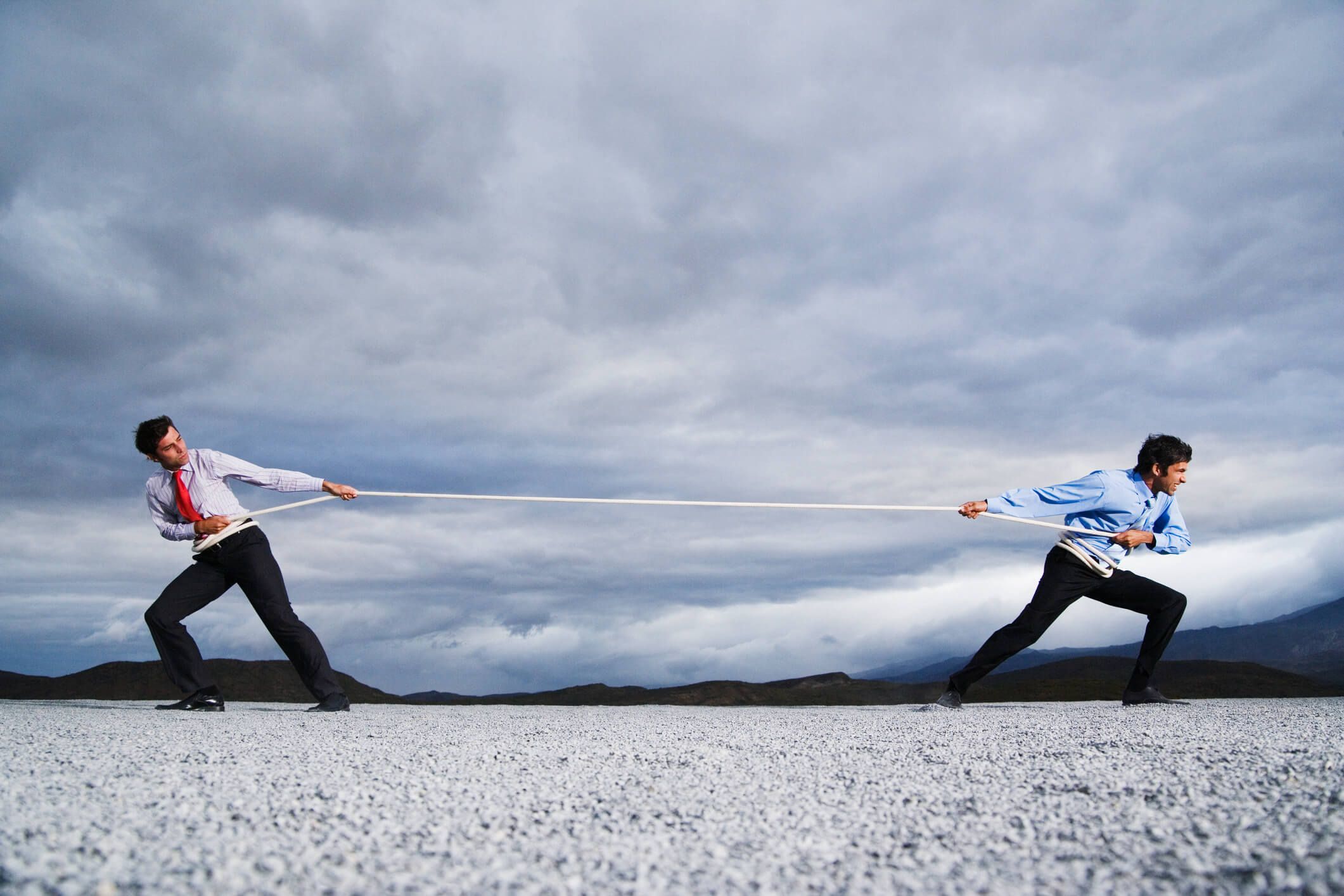 Two men stand in a desert holding a rope pulling against each other